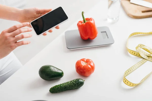 Vista recortada de niña sosteniendo teléfono inteligente con pantalla en blanco cerca de verduras, básculas y cinta métrica en la mesa de la cocina, dieta de conteo de calorías - foto de stock