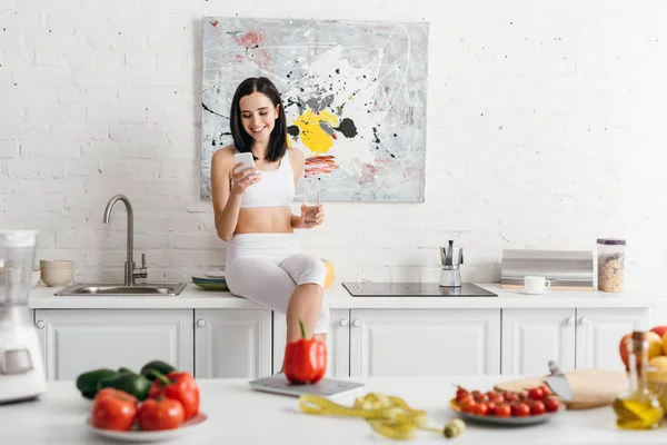 Focus selettivo di sportiva sorridente con bicchiere d'acqua e smartphone vicino alle verdure e nastro di misurazione sul tavolo della cucina, dieta per il conteggio delle calorie — Foto stock