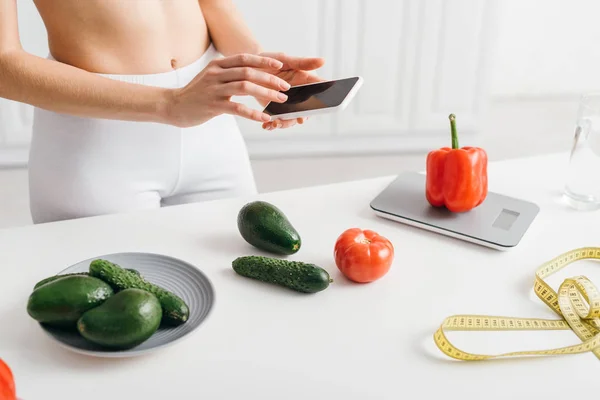 Vista ritagliata della ragazza in forma utilizzando smartphone vicino alle verdure, metro a nastro e bilancia sul tavolo, dieta conteggio calorie — Foto stock