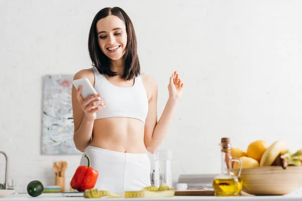 Visão de baixo ângulo da esportista sorrindo usando smartphone perto de frutas, legumes e fita métrica na cozinha — Fotografia de Stock