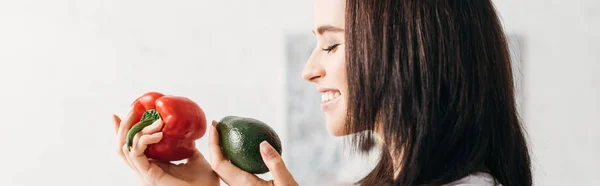 Side view of smiling girl holding bell pepper and avocado, panoramic shot — Stock Photo