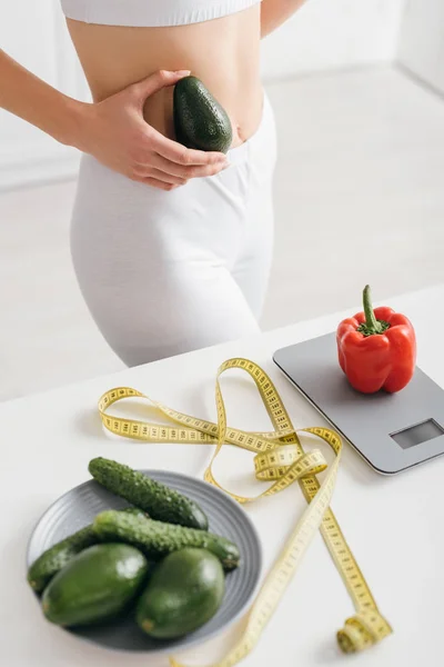 Обрізаний вид спортсменки, що тримає авокадо поблизу болгарського перцю на вагах і вимірювальної стрічки на кухонному столі, дієта підрахунку калорій — стокове фото