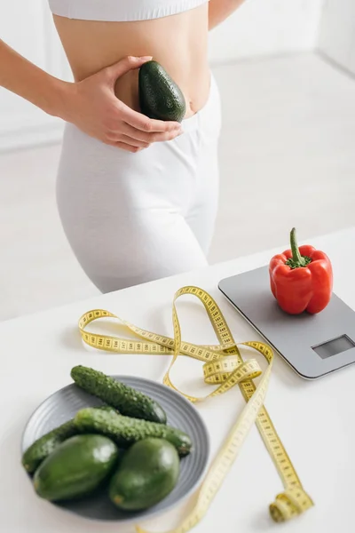 Vista cortada de esportista apto segurando abacate perto de pimentão em escalas e fita métrica na mesa da cozinha, dieta de contagem de calorias — Fotografia de Stock