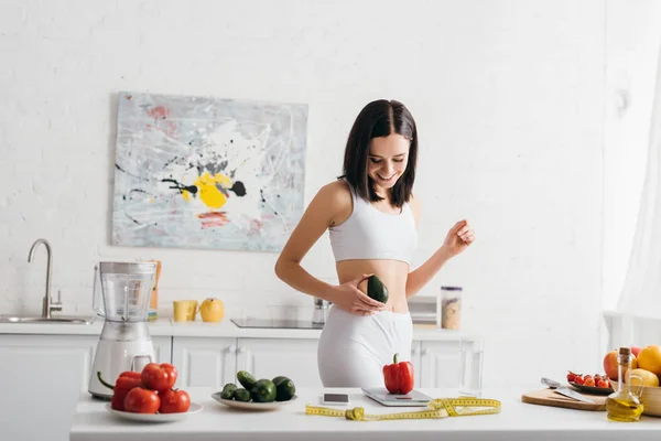 Smiling sportswoman holding avocado near fresh vegetables, scales and measuring tape on kitchen table, calorie counting diet — Stock Photo