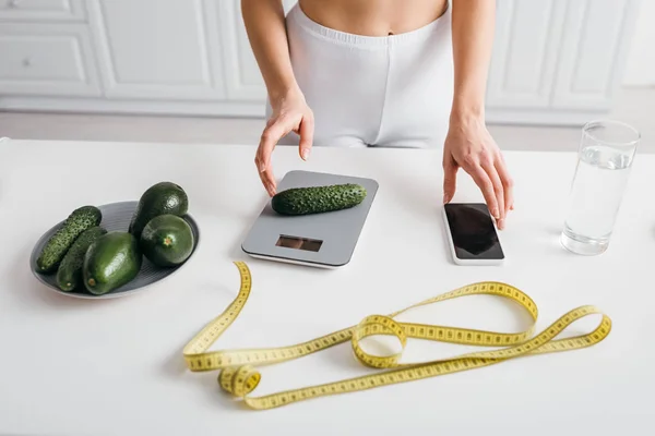 Vista cortada da mulher magra colocando pepino em escalas perto do smartphone e fita métrica na mesa da cozinha, dieta de contagem de calorias — Fotografia de Stock