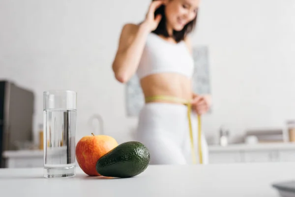 Enfoque selectivo de vaso de agua y frutas en mesa y deportista sonriente midiendo cintura con cinta adhesiva en cocina - foto de stock
