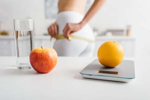 Selective focus of fruits, glass of water and scales on table near slim girl measuring hips in kitchen, calorie counting diet — Stock Photo