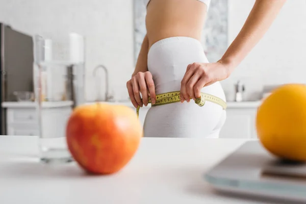 Enfoque selectivo de niña delgada midiendo caderas cerca de frutas, vaso de agua y escamas en la mesa en la cocina, dieta de conteo de calorías - foto de stock