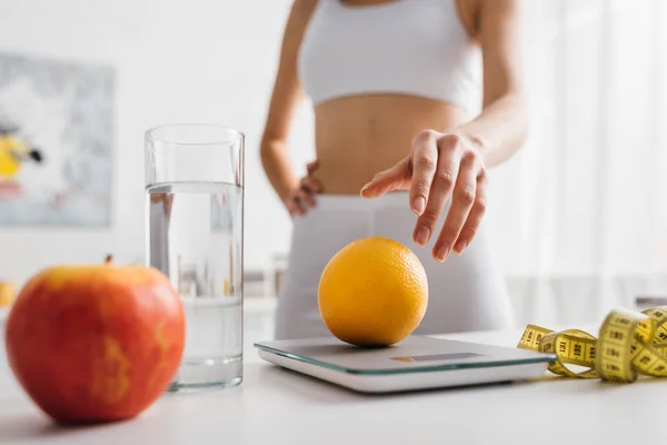 Selective focus of fit sportswoman putting orange on scales near water glass and measuring tape on kitchen table, calorie counting diet — Stock Photo