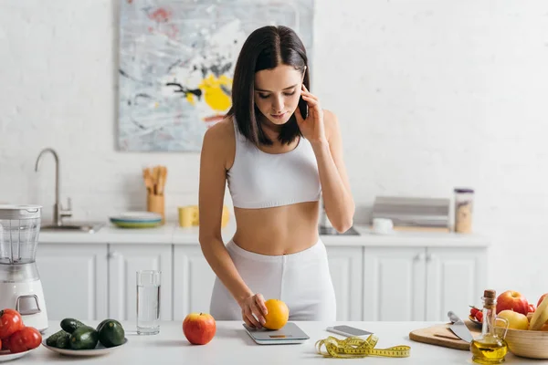 Bonita esportista pesando laranja com balanças perto de fita métrica e smartphone na mesa da cozinha, dieta de contagem de calorias — Fotografia de Stock