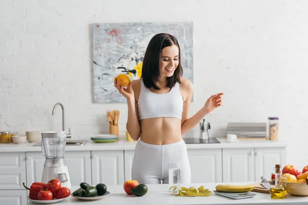 Улыбающаяся спортсменка взвешивает фрукты возле стакана воды и измеряет ленту на кухонном столе, подсчитывает калории — стоковое фото