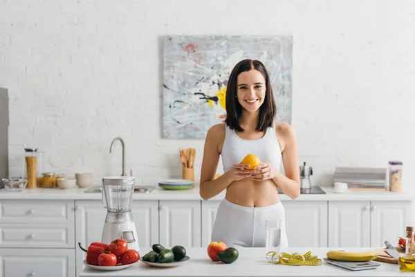 Улыбающаяся спортсменка держит апельсин возле чешуи, измеряет ленту и овощи на кухонном столе, подсчитывает калории — стоковое фото