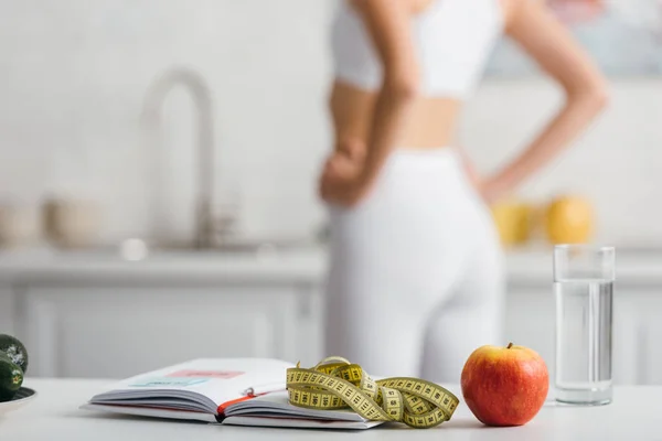 Selective focus of notebook, measuring tape and apple on table with sportswoman in kitchen — Stock Photo