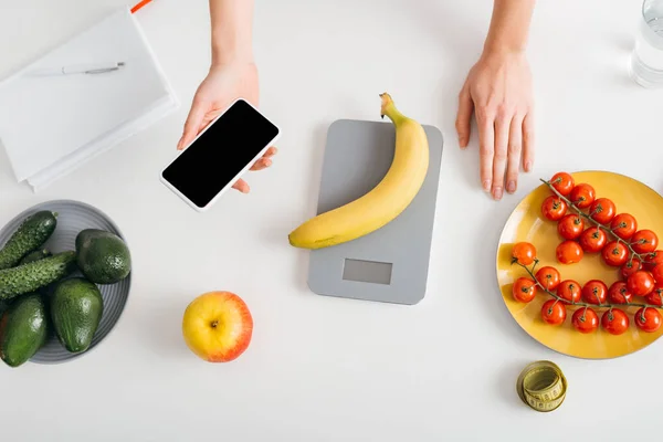 Top view of girl holding smartphone while weighing banana on kitchen table, calorie counting diet — Stock Photo
