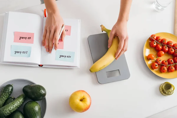 Visão superior da menina colocando banana em escalas enquanto escreve calorias no caderno, dieta de contagem de calorias — Fotografia de Stock