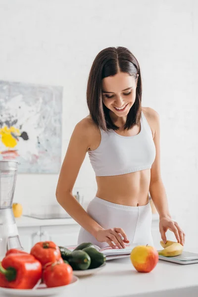 Selektiver Fokus der lächelnden Sportlerin, die Bananen wiegt und auf Notizbuch auf Küchentisch schaut, Kalorienzähler-Diät — Stockfoto