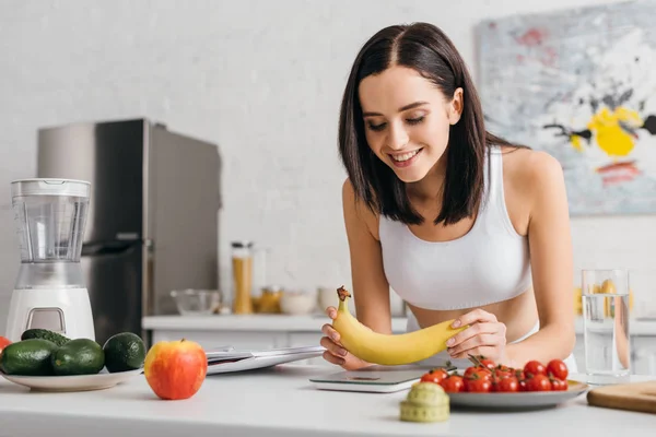 Concentration sélective de la sportive souriante mettant la banane sur des balances près du ruban à mesurer, des fruits et du carnet sur la table de cuisine, régime de comptage des calories — Photo de stock