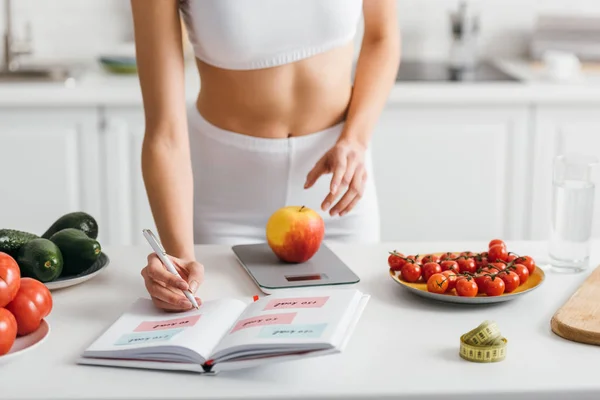 Vista recortada de la deportista en forma escribiendo calorías mientras pesa manzana en la mesa de la cocina, dieta de conteo de calorías - foto de stock