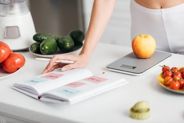 Selective focus of woman writing calories while weighing apple near fresh vegetables on kitchen table, calorie counting diet — Stock Photo