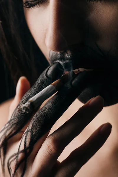 Selective focus of girl with black painted hand and lips smoking cigarette isolated on black — Stock Photo