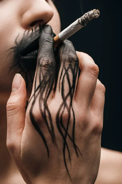 Vue recadrée de la femme avec de la peinture noire sur les lèvres et cigarette à fumer à la main isolé sur noir — Photo de stock