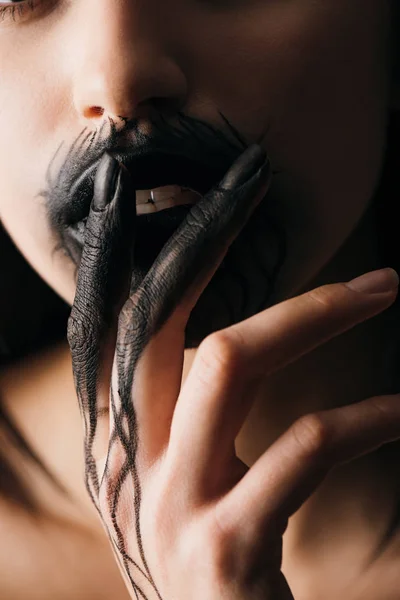 Cropped view of young woman with black paint on hand touching lips isolated on black — Stock Photo