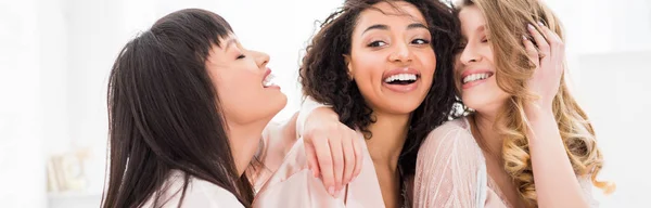 Panoramic shot of cheerful multicultural women in bathrobes hugging on bachelorette party — Stock Photo