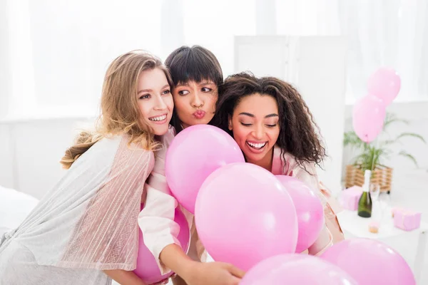 Многокультурные эмоциональные девушки веселятся с розовыми шариками в спальне — стоковое фото