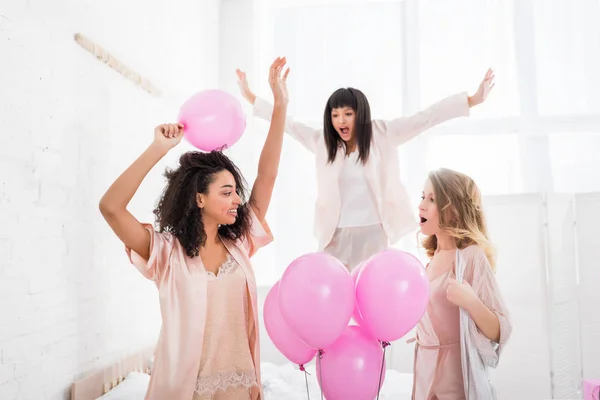 Attraktive fröhliche multikulturelle Freundinnen tanzen und springen auf dem Bett mit rosa Luftballons für Junggesellenabschied — Stockfoto