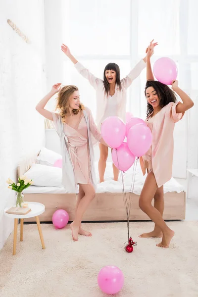 Emotional multicultural girlfriends dancing on bed with pink balloons — Stock Photo