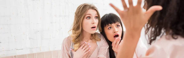 Panoramic shot of attractive shocked multicultural girls gesturing and gossiping on pajama party — Stock Photo