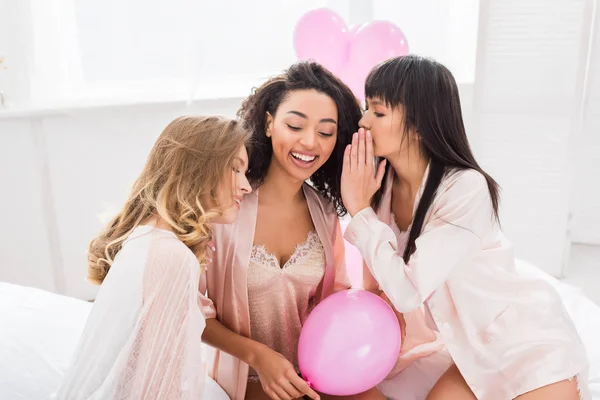Happy multicultural girlfriends whispering and gossiping on bed with pink balloons — Stock Photo