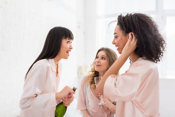 Meninas multiculturais emocionais falando ao abrir garrafa de champanhe na despedida de solteira — Fotografia de Stock