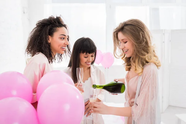 Alegre multicultural niñas verter champán de botella en vasos en despedida de soltera con globos de color rosa — Stock Photo