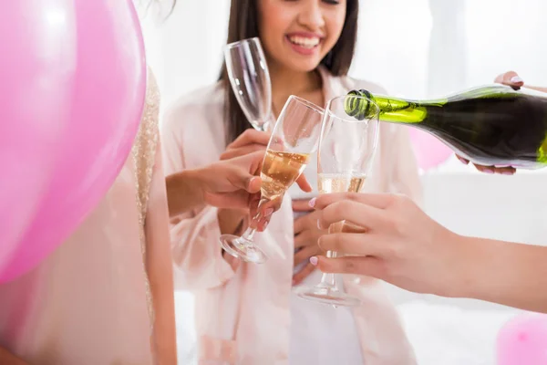 Vista recortada de chicas felices vertiendo champán de botella en vasos en despedida de soltera con globos de color rosa - foto de stock