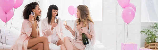 Plan panoramique de filles multiethniques heureuses avec champagne parlant sur enterrement de vie de jeune fille avec ballons — Photo de stock