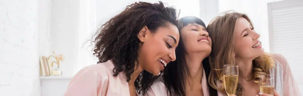 Panoramic shot of happy multicultural girlfriends having fun and holding champagne glasses on pajama party — Stock Photo