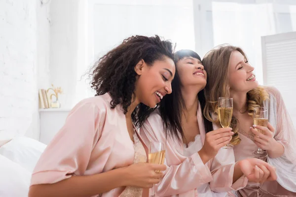 Lächelnde multiethnische Freundinnen, die Spaß haben und Champagnergläser auf Pyjama-Party halten — Stockfoto