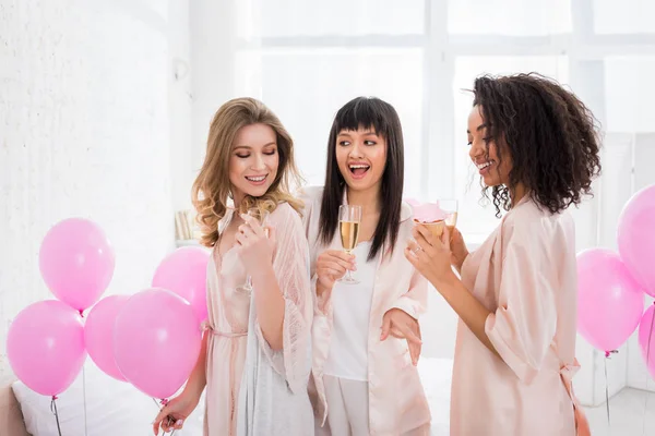 Счастливые мультиэтнические девушки веселятся с шампанским и кексом во время девичника с розовыми шариками — стоковое фото