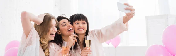 Plano panorámico de chicas emocionales multiculturales con copas de champán tomando selfie en el teléfono inteligente durante la fiesta de pijama - foto de stock