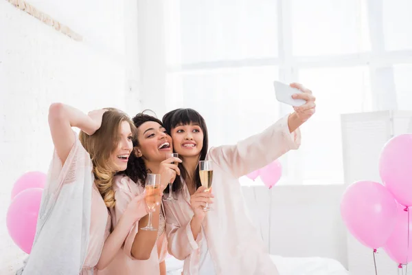 Chicas multiculturales atractivas con copas de champán tomando selfie en el teléfono inteligente durante la fiesta de pijama - foto de stock