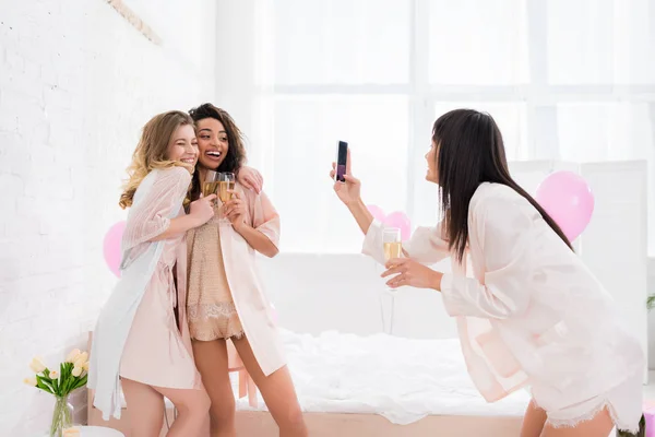 Beautiful girl taking photo of multicultural happy girlfriends with glasses of champagne on smartphone during pajama party — Stock Photo