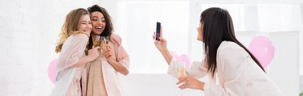 Plano panorámico de chica alegre tomando foto de novias multiculturales con copas de champán en smartphone durante la fiesta de pijama - foto de stock