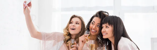 Plano panorámico de chicas multiétnicas sonrientes con copas de champán tomando selfie en el teléfono inteligente durante la fiesta de pijama - foto de stock