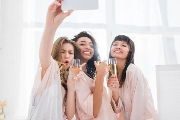 Emotional multiethnic girlfriends with glasses of champagne taking selfie on smartphone during pajama party — Stock Photo