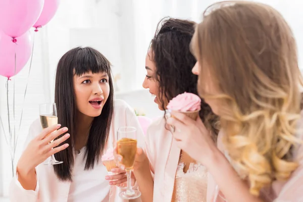Chicas multiculturales sorprendidas divirtiéndose con champán y magdalena durante la despedida de soltera con globos rosados - foto de stock