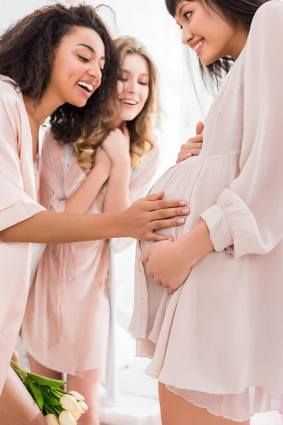 Многокультурные девушки касаются живота молодой беременной женщины во время девичника — стоковое фото