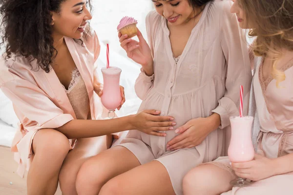 Smiling multicultural girlfriends and young pregnant woman holding cupcake and glasses with milkshakes on baby shower — Stock Photo