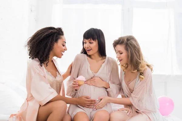 Novias multiculturales tocando barriga de mujer embarazada joven sosteniendo cupcake rosa en baby shower - foto de stock
