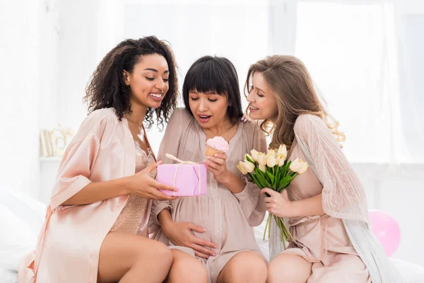 Novias multiculturales y joven embarazada sosteniendo cupcake, flores y regalo en baby shower - foto de stock